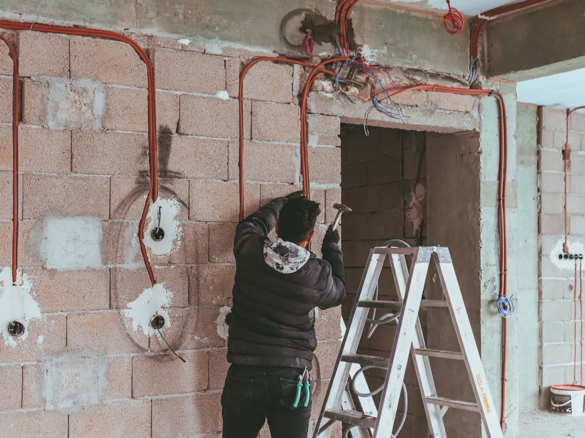 Électricien sur un escabeau dans une pièce en travaux, gaines électriques sorties des murs